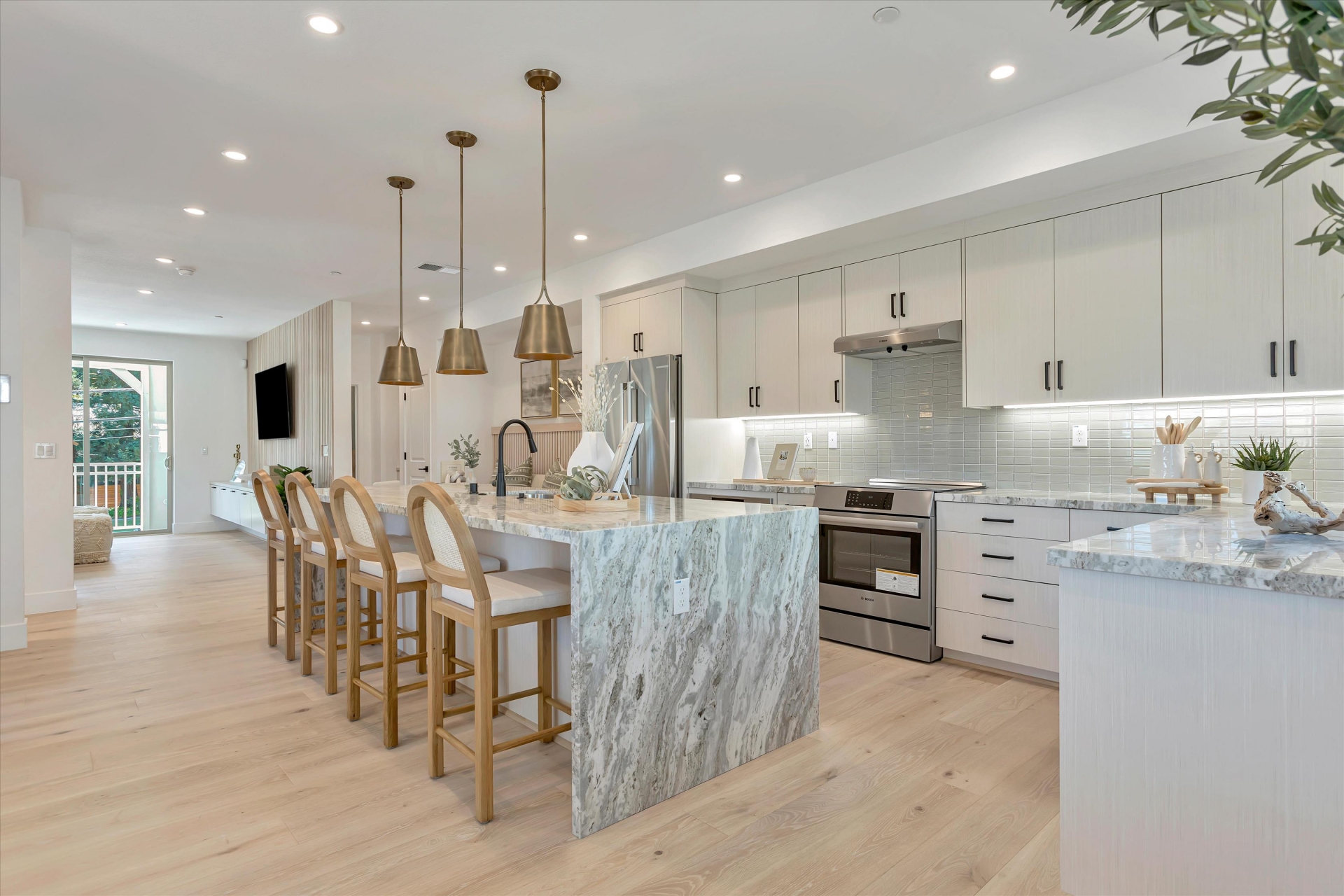 Bright modern kitchen at Oak Grove in Walnut Creek featuring marble waterfall island, brass pendant lighting, light wood flooring, and sleek cabinetry with stainless steel appliances.